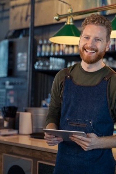 Successful small business owner using digital tablet and looking at camera. Happy smiling waiter wearing apron and holding digital tablet ready to take order. Portrait of young entrepreneur of coffee shop standing at counter with copy space.