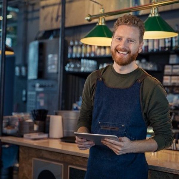 Successful small business owner using digital tablet and looking at camera. Happy smiling waiter wearing apron and holding digital tablet ready to take order. Portrait of young entrepreneur of coffee shop standing at counter with copy space.
